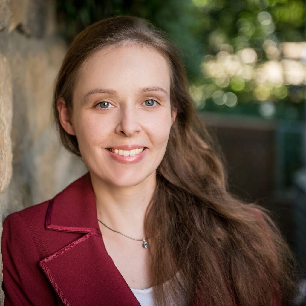 Autorinnenfoto Anika Herber. Junge lächelnde Frau mit langem brauen Haar und rotem Blazer